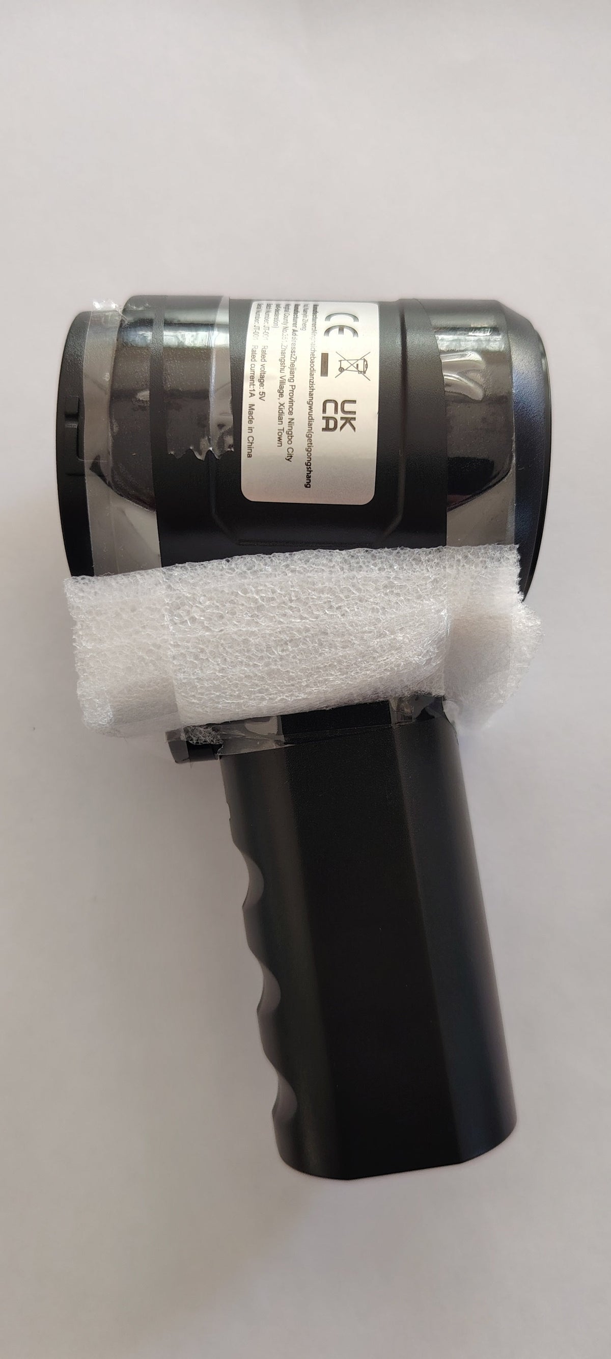 A black handheld electronic device with a protective white foam sleeve around its middle, photographed against a plain off-white background.